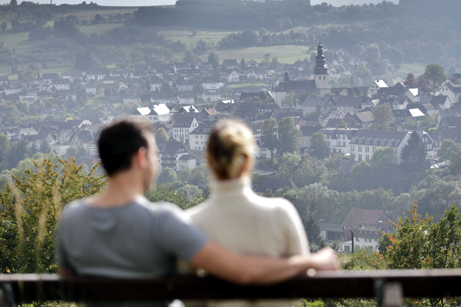 Ein Paar sitzt auf einer Bank am Wanderweg und Blickt auf das Fachwerkstädchen Hallenberg bei klarem Sommerwetter