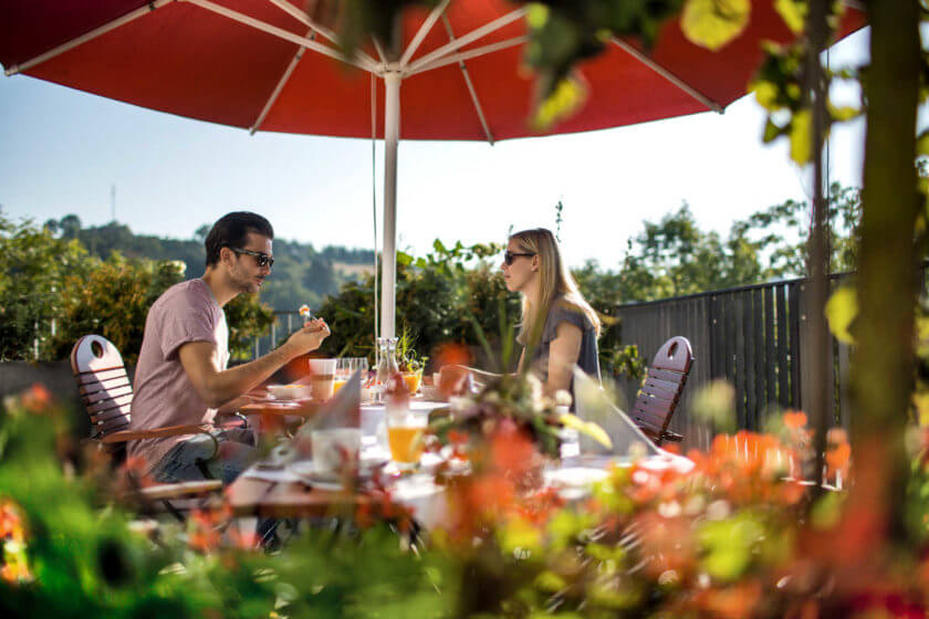 Ein Paar frühstückt bei bestem Sommerwetter auf der Terrasse unter einem roten Sonnenschirm