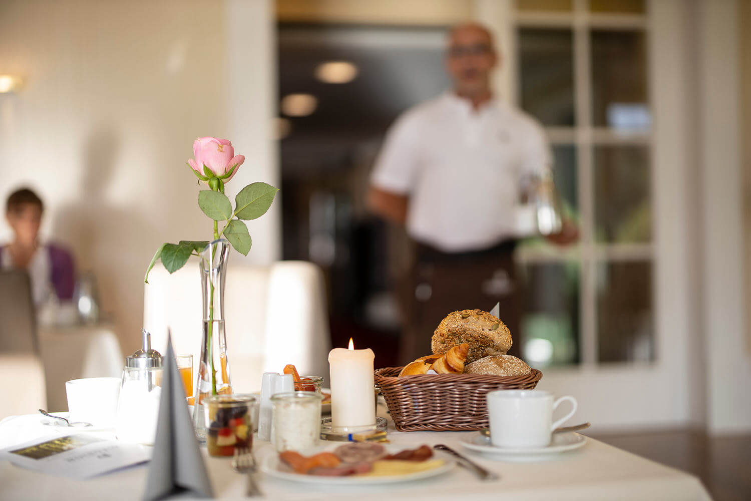 Im Vordergrund ein reichlich gefüllter Frühstückstisch, während im Hintergrund Kaffee gebacht wird - Hotel Diedrich