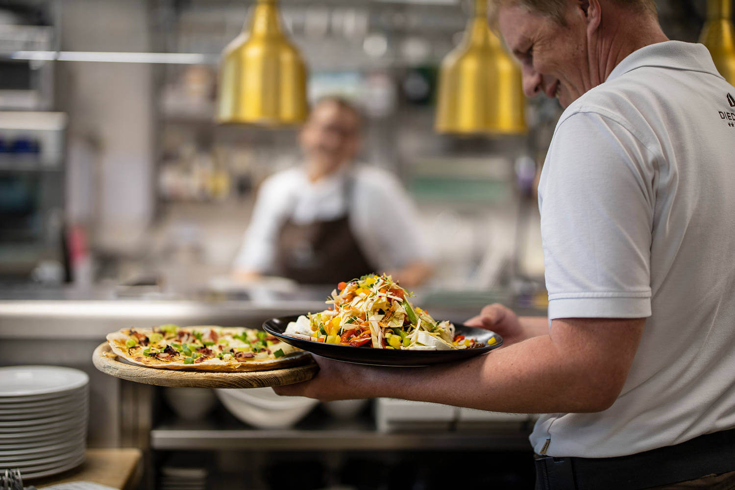 Ein Servicemitarbeiter stellt auf seinen linken Arm einen Flammkuchen und einen Rohkostsalat zum Servieren - Hotel Diedrich