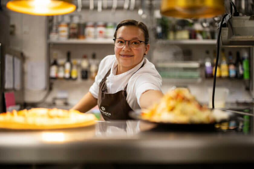 A cook smiles into the camera while placing a plate on the kitchen pass - Hotel Diedrich