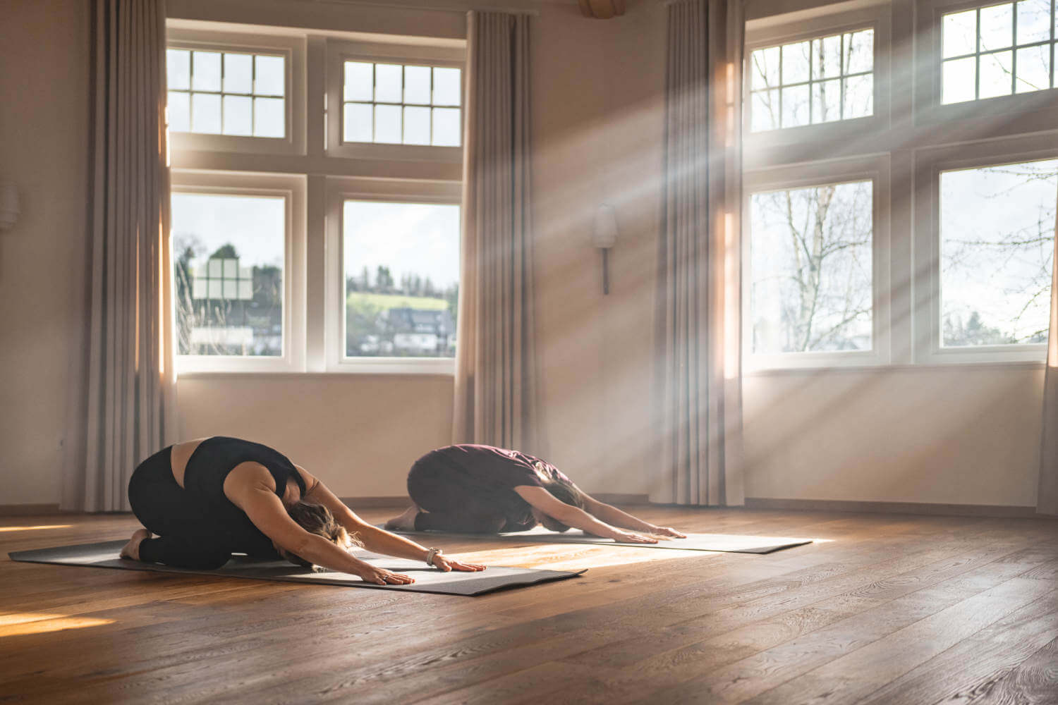 Zwei Frauen knien auf ihren Yogamatten und strecken ihre Arme nach vorn, das Sonnenlicht scheint in den Yogaraum