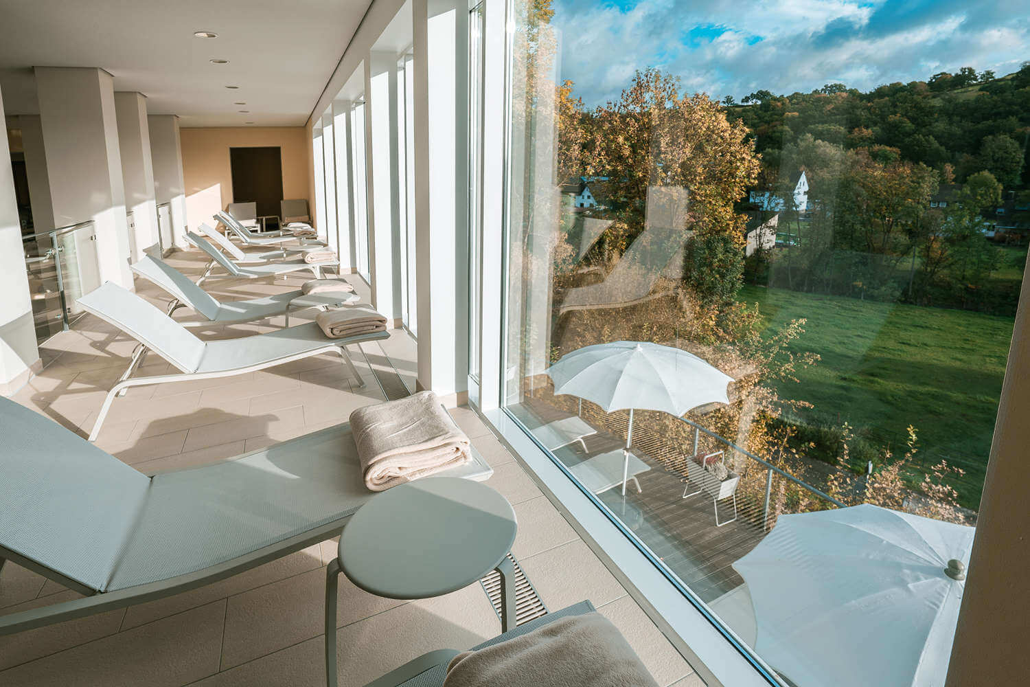 View from the upper floor of the pillar bath through the panoramic windows to the outside, with loungers lined up in front of it - Wellnesshotel Sauerland