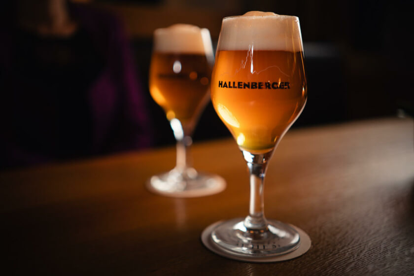  Zwei Gläser bernsteinfarbenes Hallenberger Bier stehen auf einem Eichenholztisch.