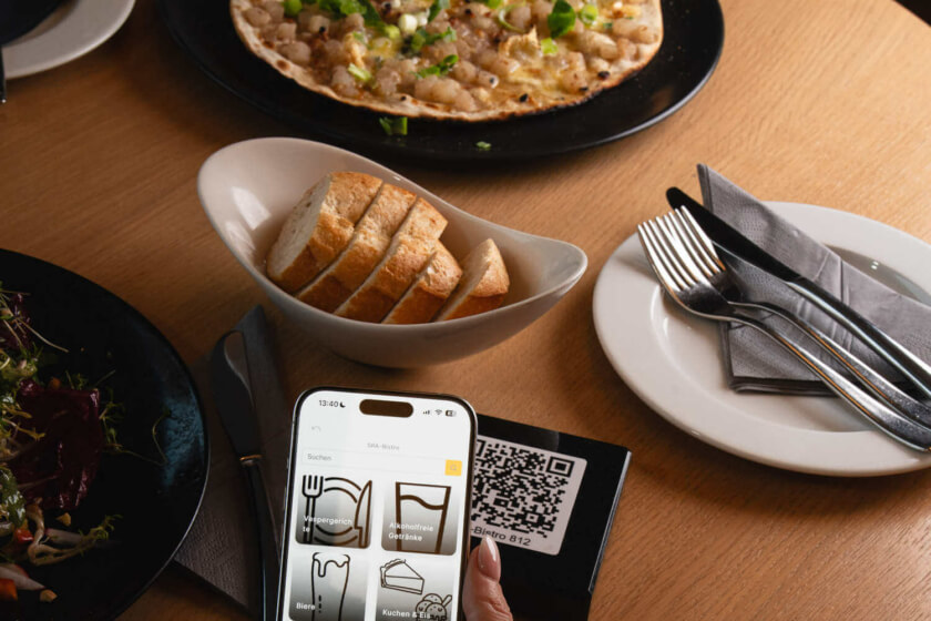 Eine Hand hält ein Smartphone mit geöffneter Speisekarten-App über einem Tisch mit leckerem Essen und einem QR-Code.
