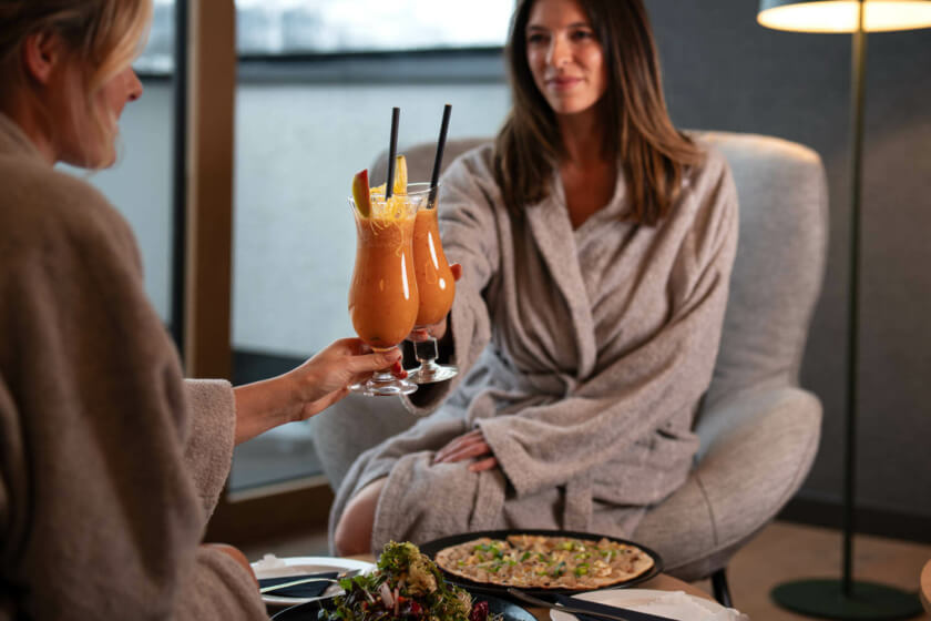 Two women in bathrobes enjoy a healthy smoothie together while sitting in a cozy wellness bistro with natural light.