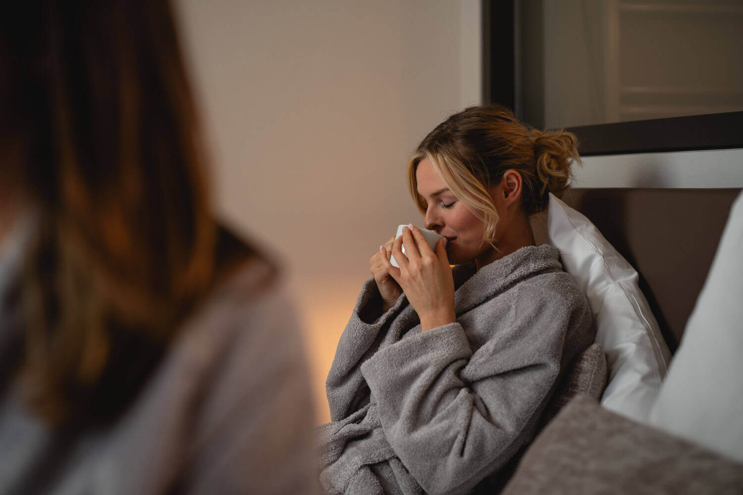 Eine Frau im Bademantel sitzt auf ihrem Bett und genießt den Duft ihrer Tasse Tee, die sie sich mit beiden Händen an den Mund hält.