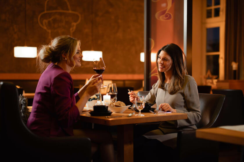 Zwei elegante Frauen stoßen mit Wein an und genießen eine Mahlzeit in einem Restaurant mit gemütlicher Atmosphäre.