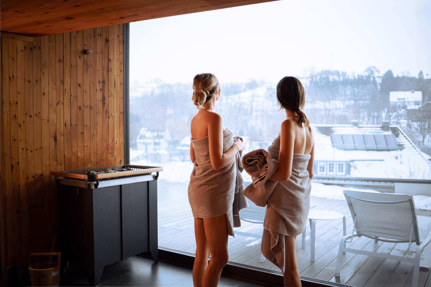 Zwei Frauen in Bademänteln stehen vor einer großen Fensterfront und blicken auf eine verschneite Landschaft hinaus.