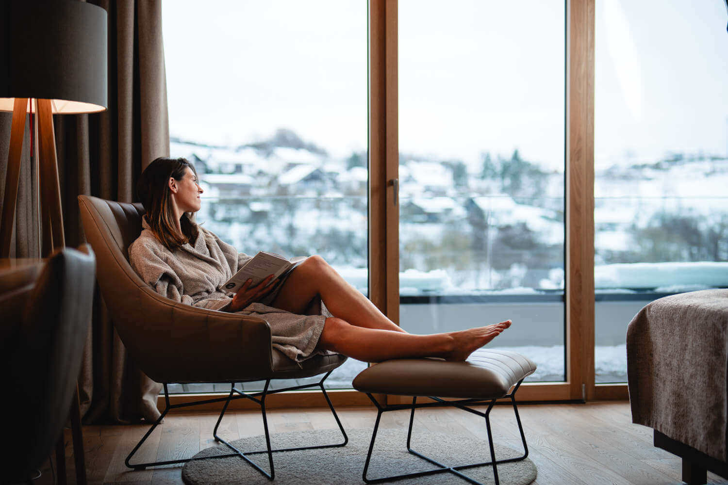 Eine Frau sitzt in Ihrem Zimmer mit den Beinen hoch auf dem Sessel und schaut verträumt nach draußen auf die Winterlandschaft.