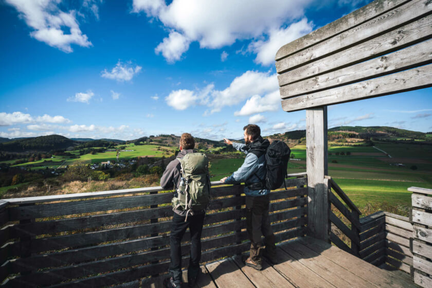 Zwei Wanderfreunde stehen auf dem Sauerland Stabilstuhl und zeigen in die hügelige Landschaft